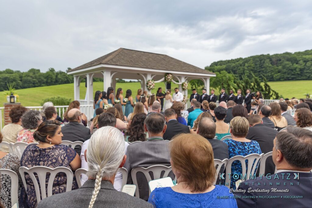 Outdoor wedding ceremony with guests seated, a bridal party at the front, and a gazebo in the background, surrounded by greenery.