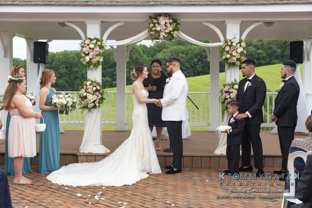 A couple stands at an outdoor wedding altar exchanging vows. Bridesmaids and groomsmen are on either side, with a child holding a sign. The altar is decorated with flowers.