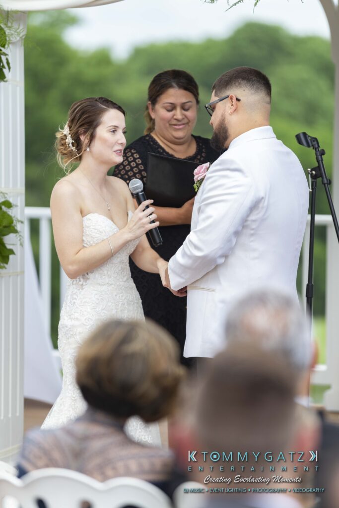 A bride and groom in a white wedding attire hold hands during a ceremony. The bride is speaking into a microphone. A woman stands behind them, smiling, with a bouquet in her hands.