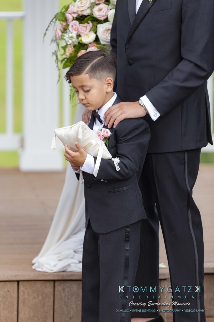 A young boy in a suit holds a pillow, standing next to an adult on a wooden platform. A bouquet of roses is visible in the background.