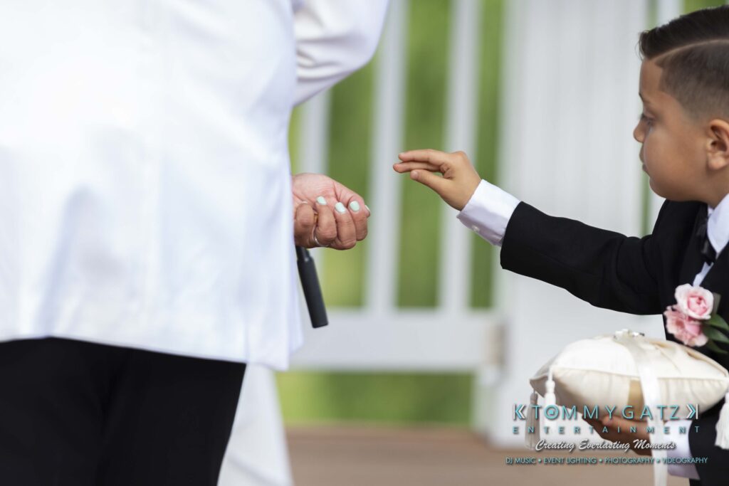 An adult holds a ring as a child in a suit reaches towards it. The child carries a pillow with rings. The background features white railings.