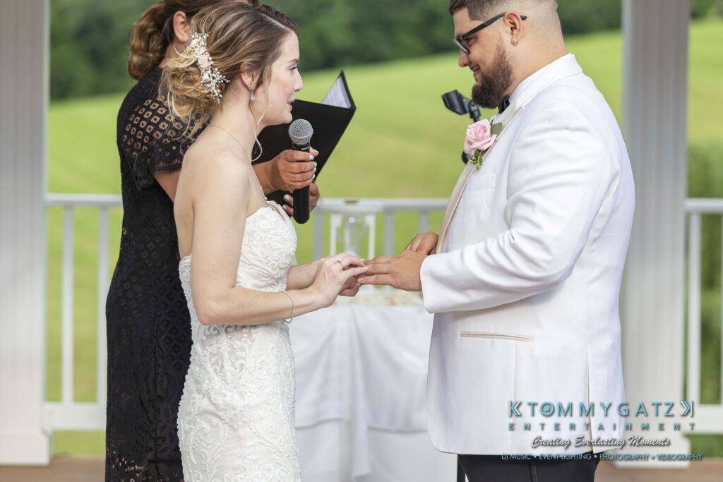 A couple exchanges rings during their wedding ceremony. The officiant reads from a script, and a guest watches. The event takes place outdoors with greenery in the background.