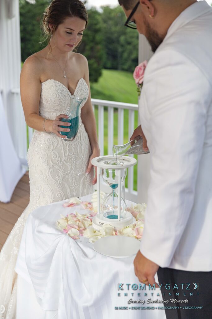 A couple conducts a sand ceremony at their wedding. The bride holds a blue sand vase, while the groom pours sand into a shared container. They stand on a porch with a green landscape in the background.