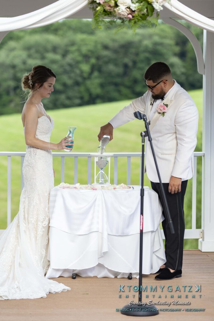 A bride and groom pour sand into an hourglass during a wedding ceremony. They stand on a gazebo with a scenic background.