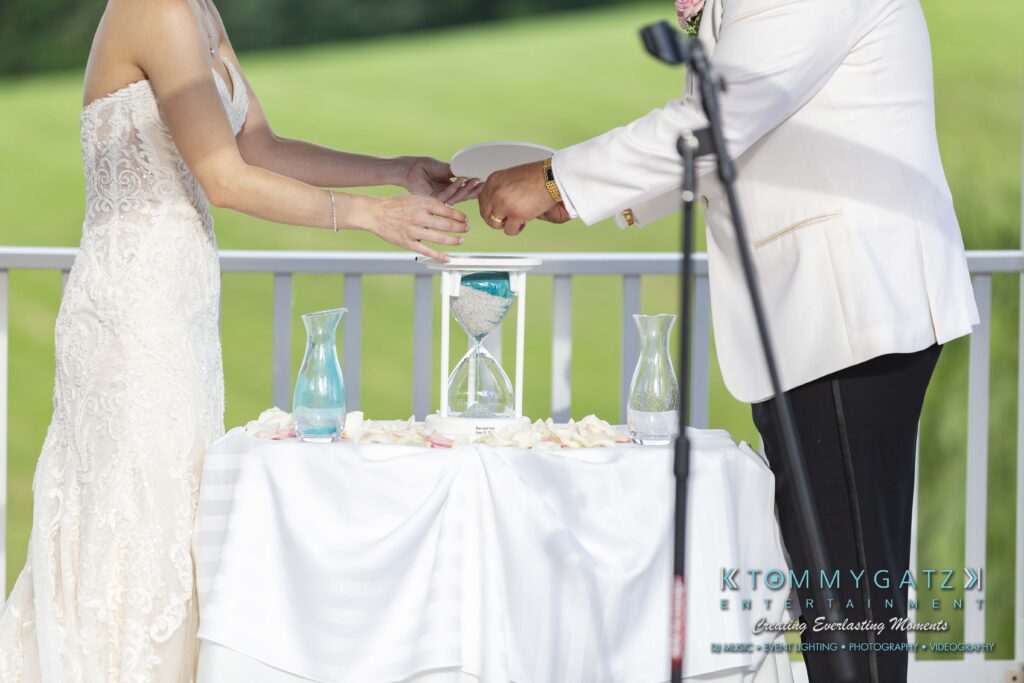 A bride and groom pour sand into an hourglass during a wedding ceremony, standing behind a table with a white cloth and rose petals.