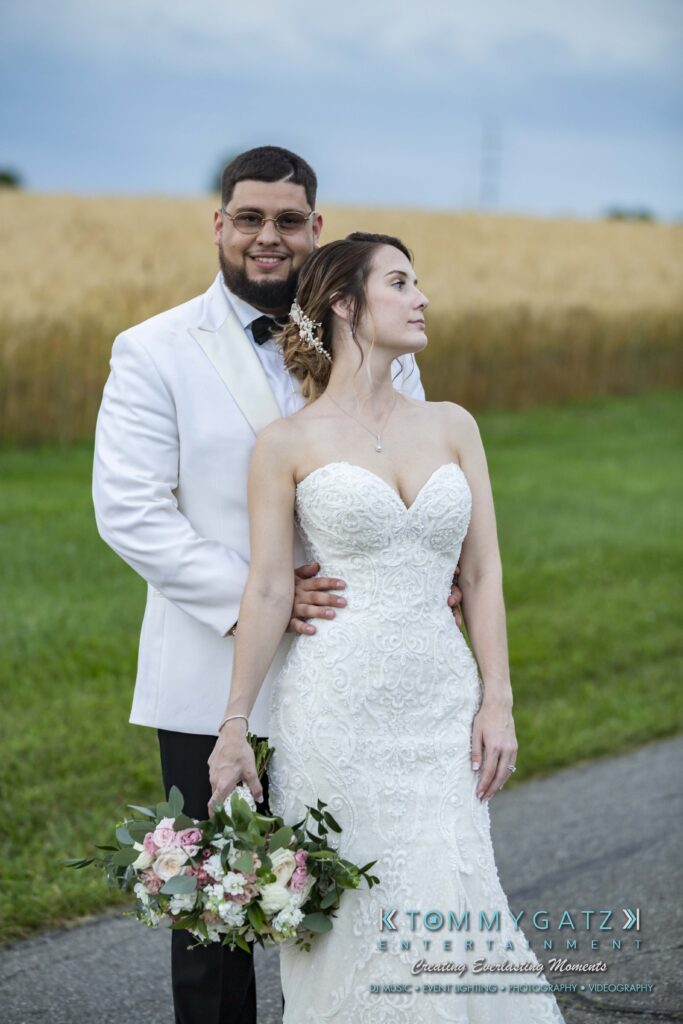 A bride and groom pose outdoors, with the groom in a white jacket and the bride in a white gown holding a bouquet. They stand on a path next to a grassy field.