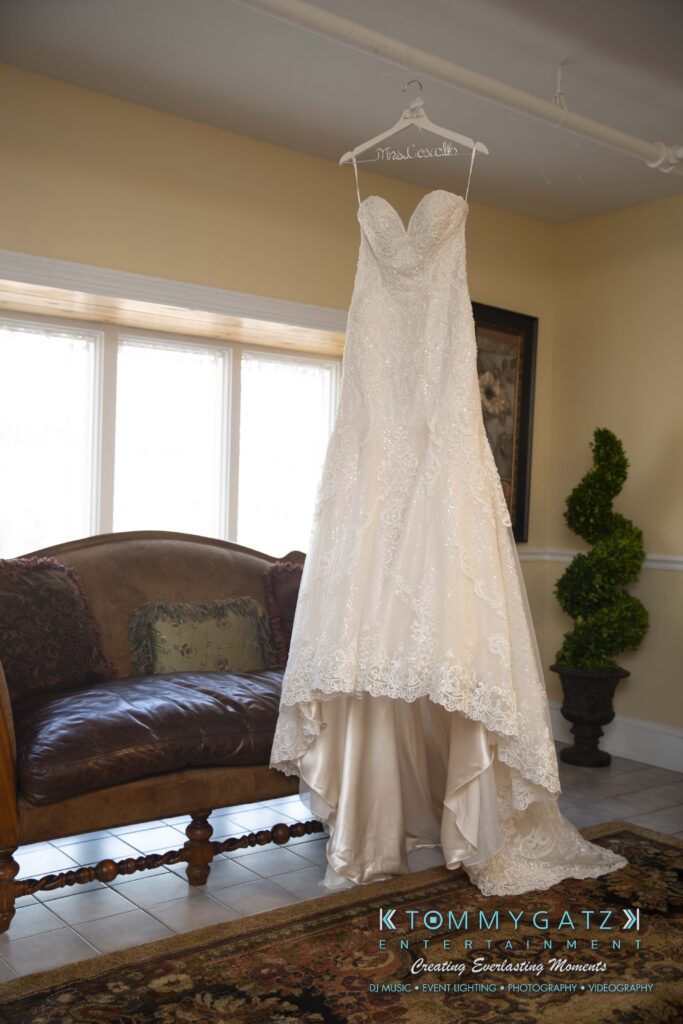 A white lace wedding dress hangs on a hanger in a room with a sofa and plants.