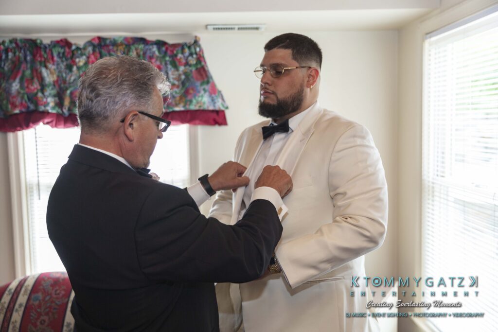 Two men in formal attire stand indoors. One helps adjust the other’s white suit jacket. A colorful curtain and a logo for Tommy Gatz Entertainment are visible.