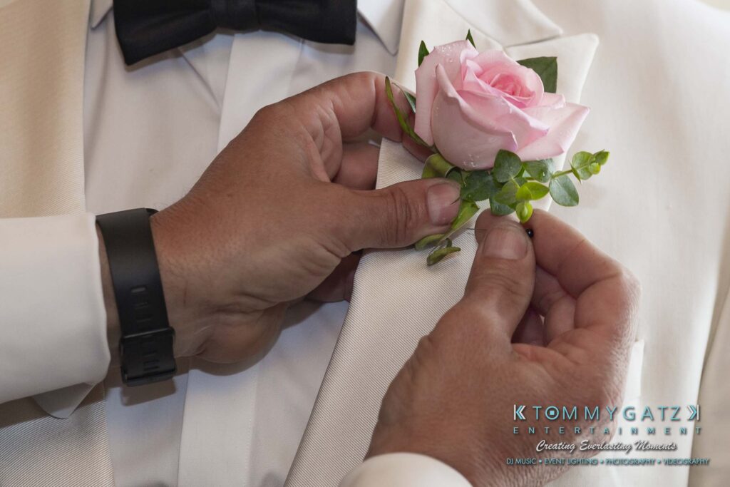 Person pinning a pink rose boutonniere on a white suit jacket, with visible hands and a wristwatch, alongside Tommy Gatz Entertainment branding.