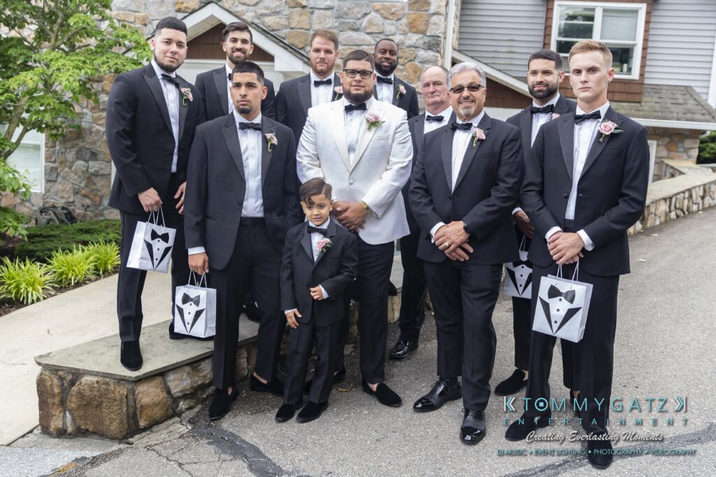 A groom in a white tuxedo stands with groomsmen and a child, all in black suits with bow ties, holding tuxedo-themed gift bags, in an outdoor setting near a stone building.