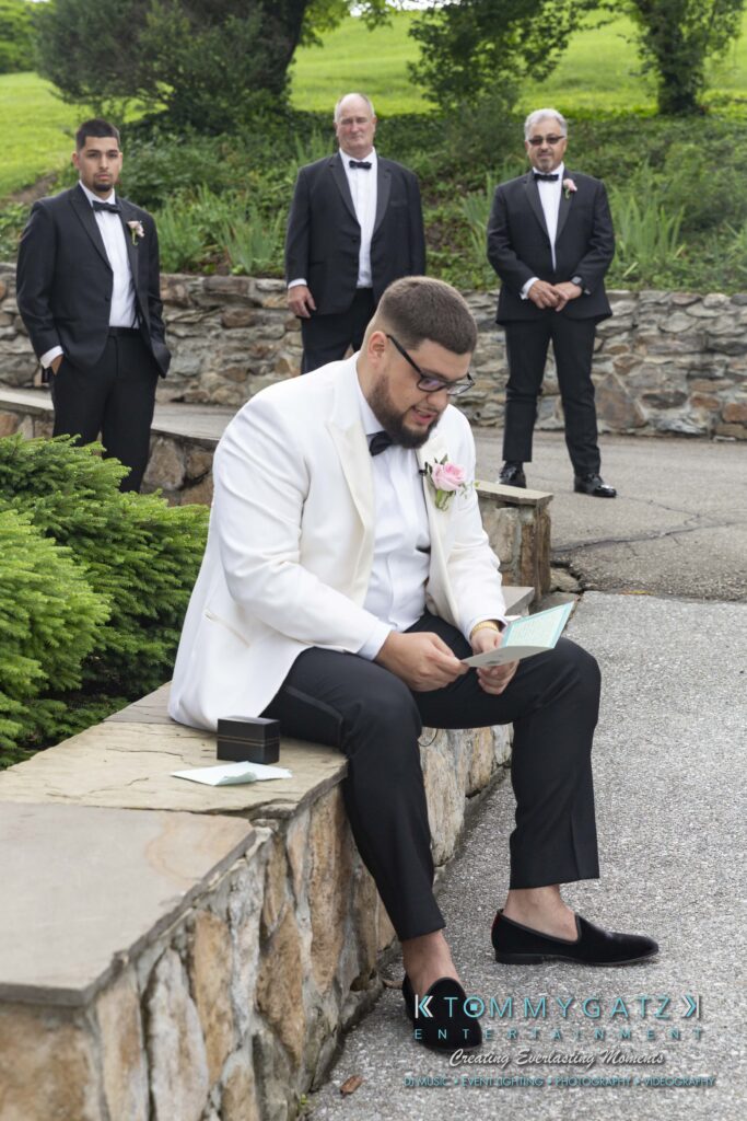 A man in a white jacket reads a card while sitting on a stone ledge. Three men in black suits stand in the background. A small black box is on the ledge beside him.