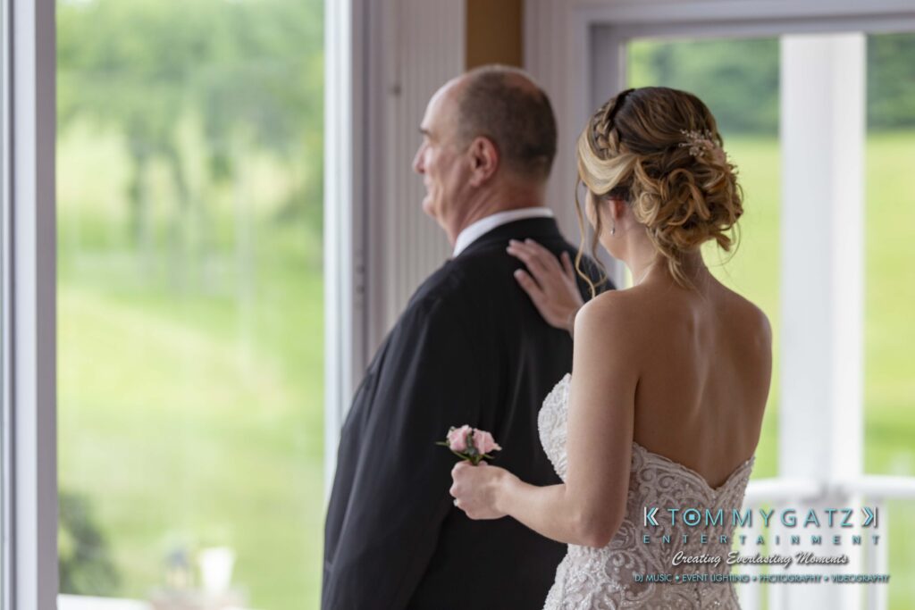 Bride in a white dress with a pink corsage places her hand on an older man's shoulder as they look out a window.