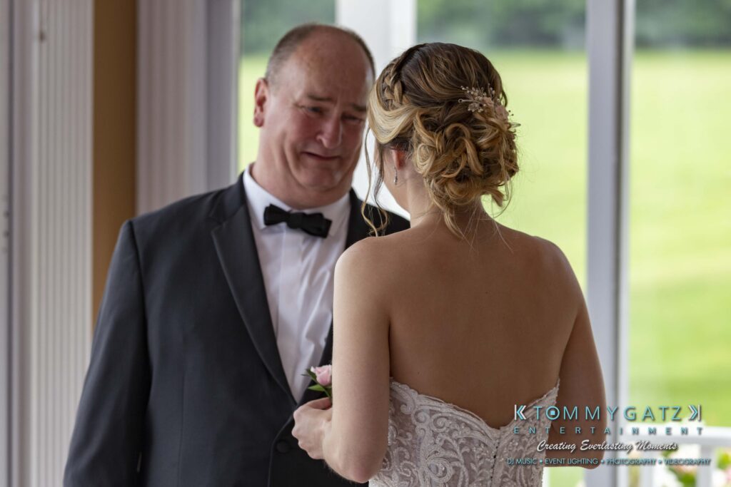 A man in a tuxedo and a woman in a white dress stand indoors. The man appears emotional.