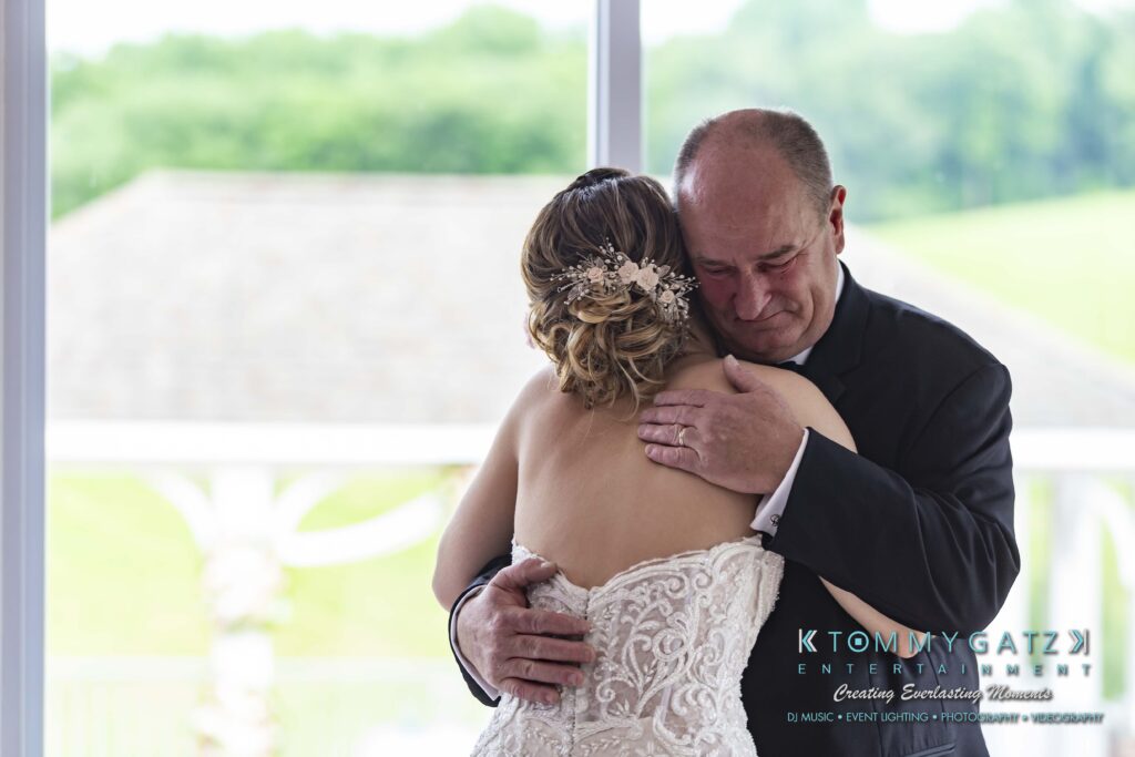 A father embraces his daughter in a tender moment at her wedding, facing a window with a blurred garden view.