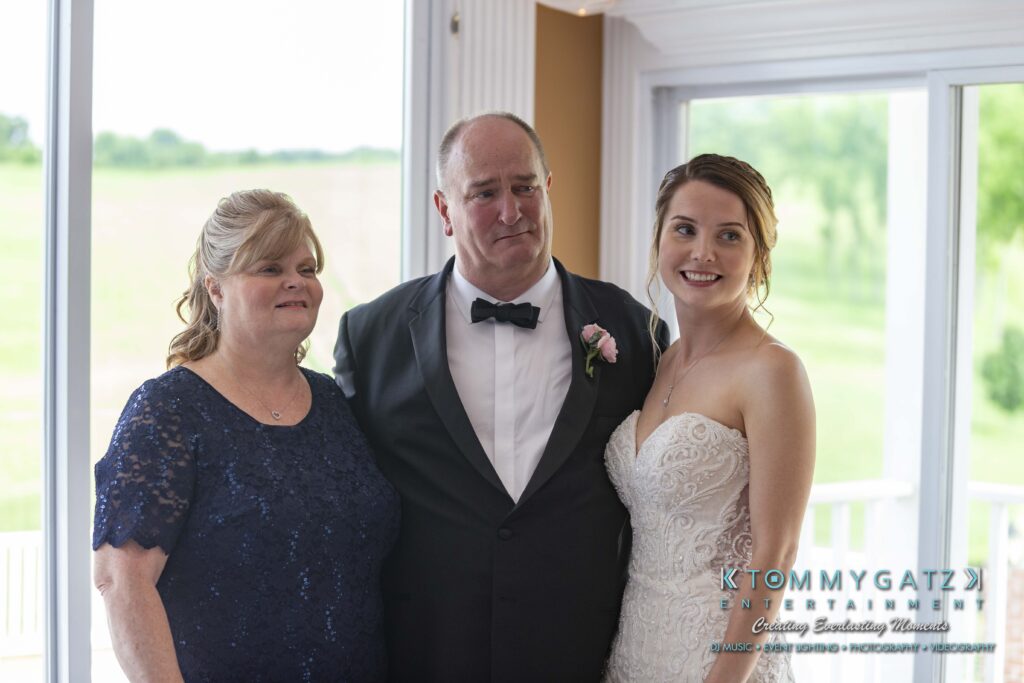 Three people are posing inside near a window. The man in a black suit stands between two women, one in a navy dress and the other in a white dress.