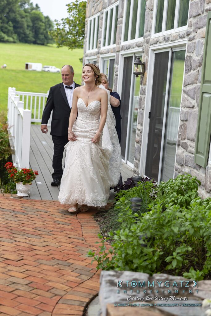 Bride walking down a brick pathway outside next to a stone building, accompanied by two people.
