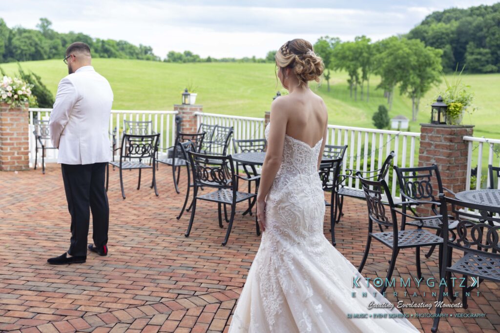 Bride in a lace gown approaches a man in a white jacket on a brick patio overlooking a grassy landscape.