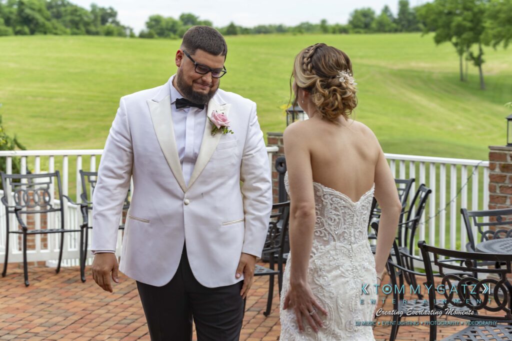 A couple stands on a patio. The man is wearing a white suit with a pink boutonniere, and the woman is in a strapless white dress. They appear to be outdoors with a grassy field in the background.