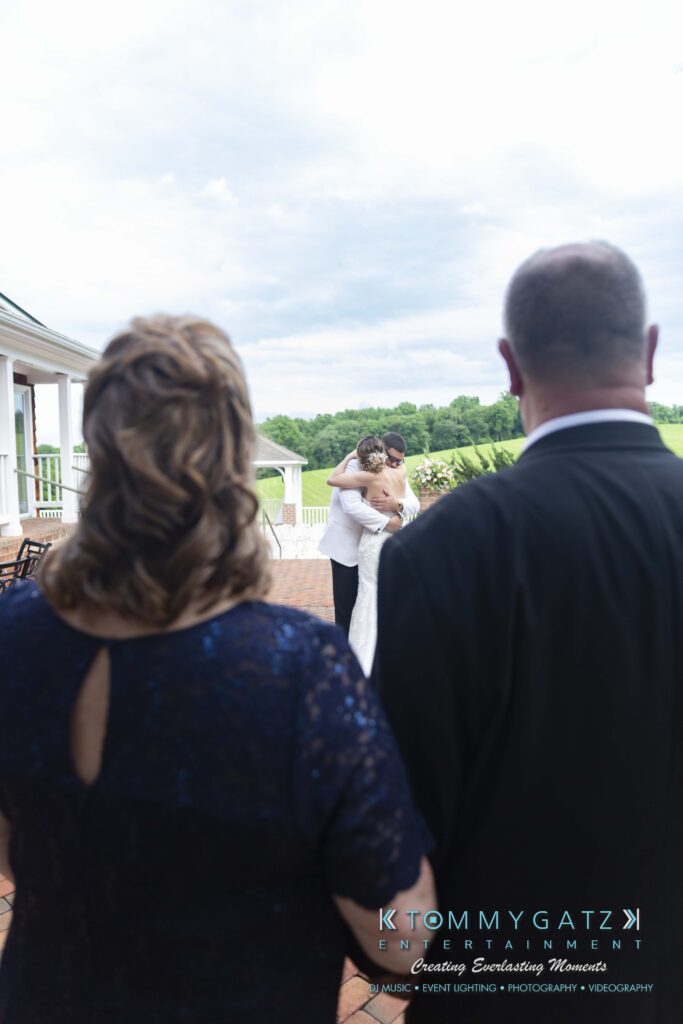 Two people embrace on a patio overlooking a scenic landscape, watched by a man and woman from behind.
