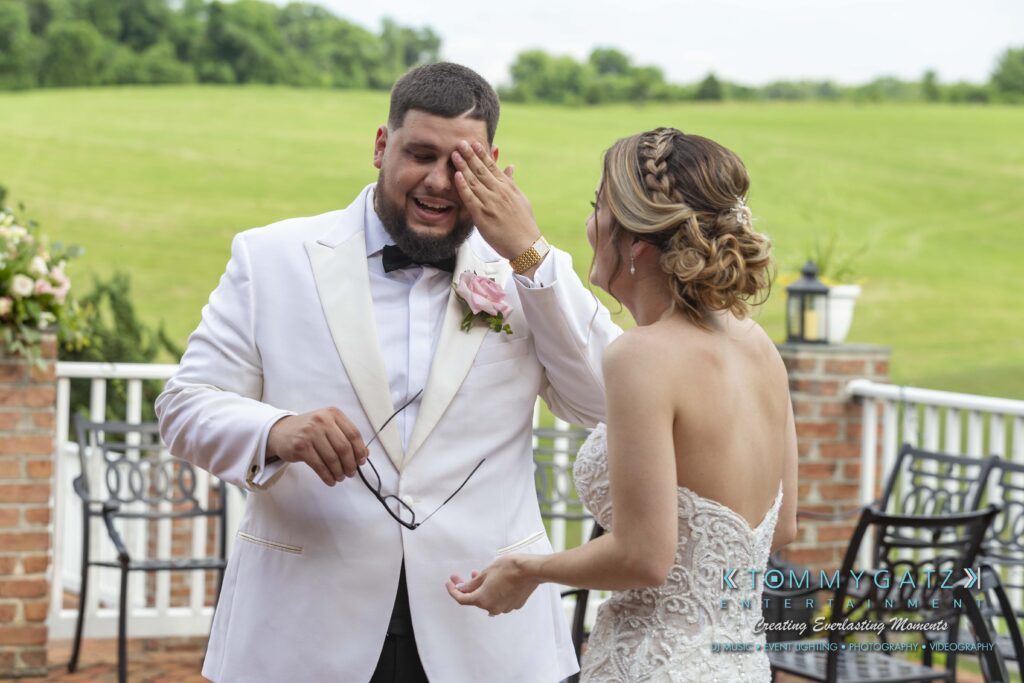 A groom in a white suit wipes his face while holding glasses. The bride in a white dress smiles and touches his cheek. They stand on a patio with a green field in the background.