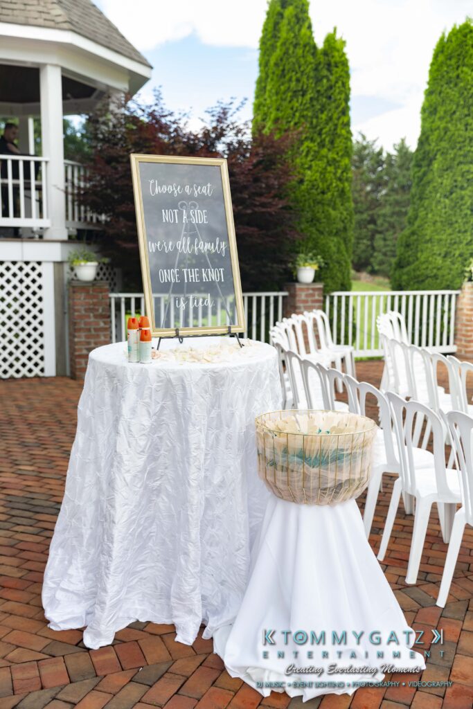 Outdoor wedding setup with a sign on a small round table reading "Choose a seat, not a side, we're all family once the knot is tied." A basket is beside the table, with white chairs arranged in rows.