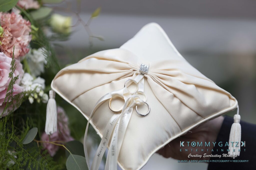 Ivory ring bearer pillow with ribbon and rings, set against a backdrop of pink and white flowers and greenery.