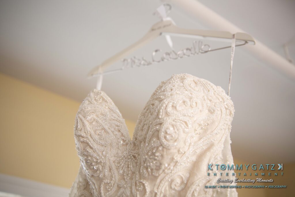 Close-up of an intricately beaded white wedding dress on a hanger, with "Mrs." on the hanger. Logos and text visible at the bottom right corner.