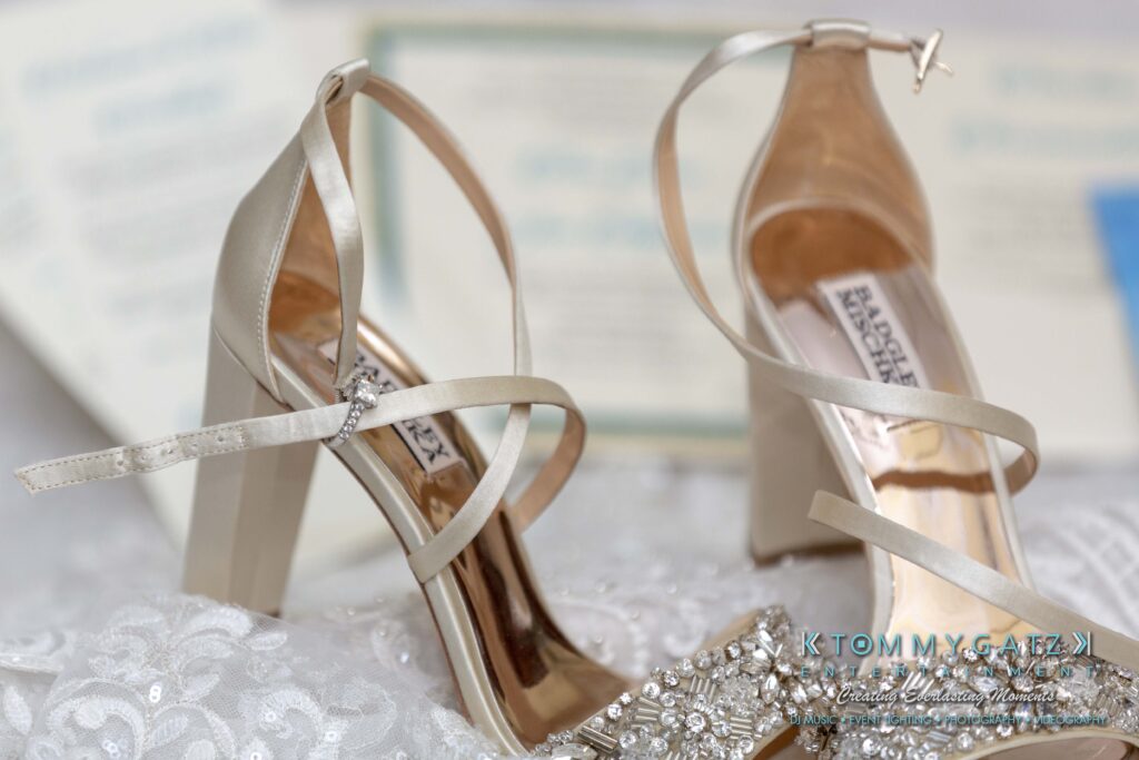 Elegant strappy high heels with rhinestone details placed on white lace fabric, invitation card blurred in background.