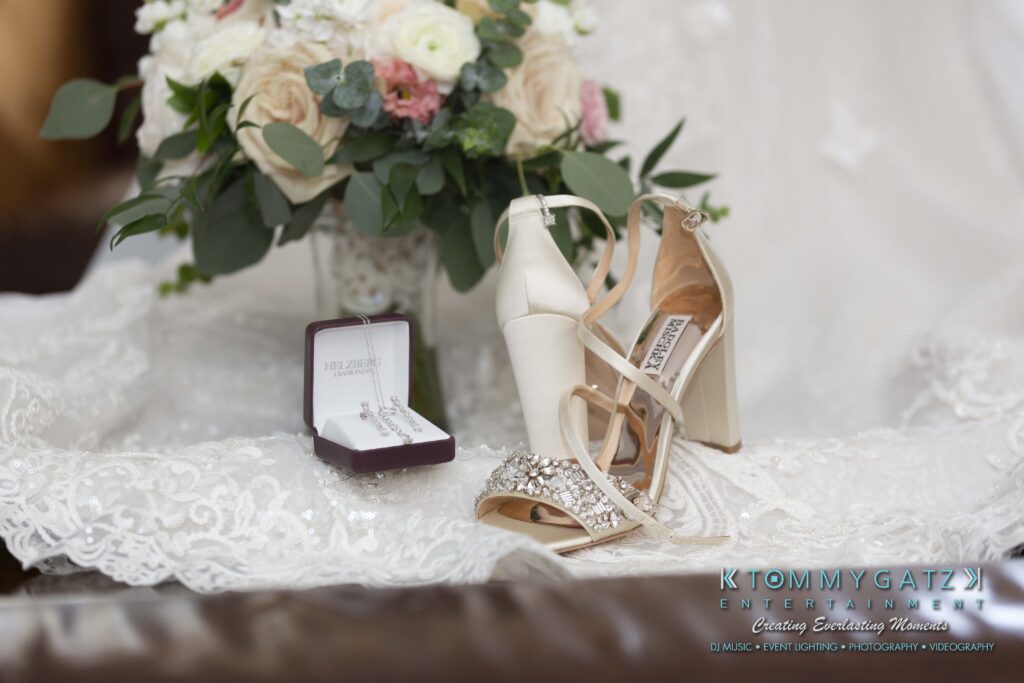 White high-heeled wedding shoes on lace fabric; next to them is an open jewelry box containing a necklace. In the background, a bouquet of white and pink flowers.