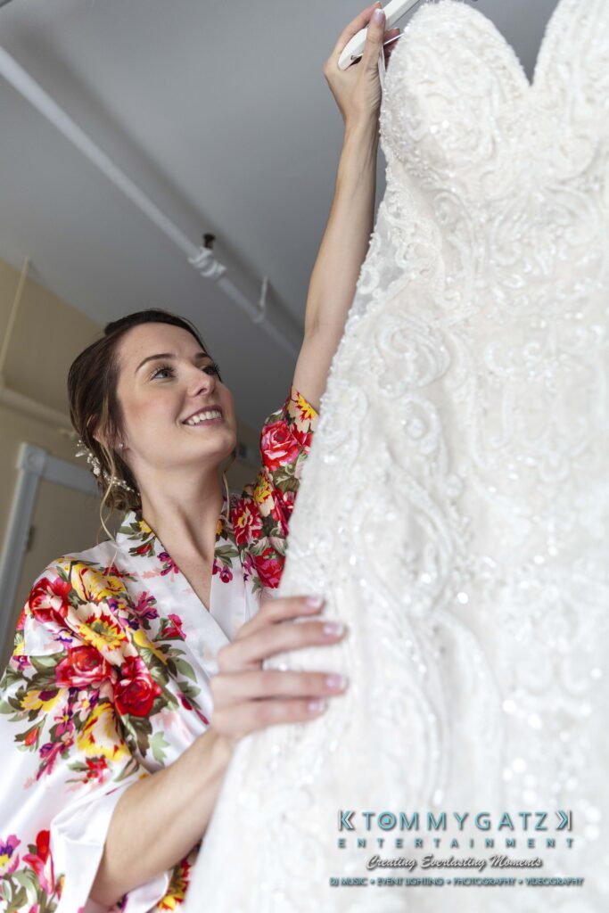 A woman in a floral robe smiles while holding a white wedding dress on a hanger.