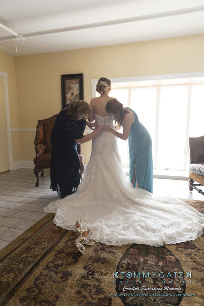 Two women help a bride adjust her lace wedding dress in a sunlit room. The bride stands on a patterned rug, and a pair of heeled shoes is on the floor.