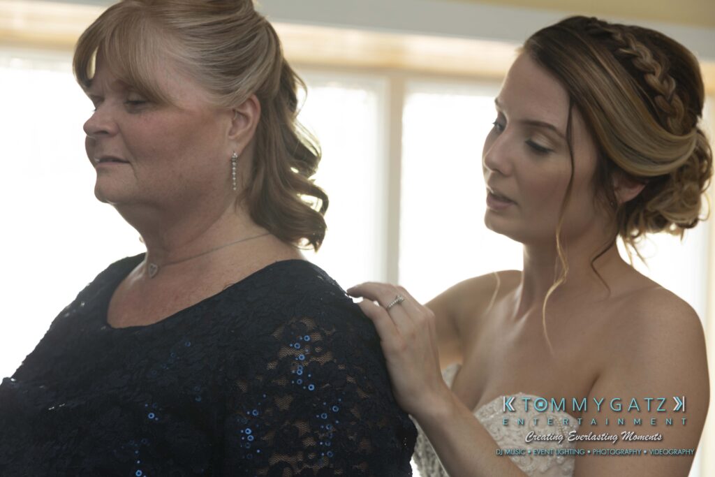A bride assists a woman in a dark dress, adjusting the shoulder area. They are indoors, with natural light shining through the window behind them.
