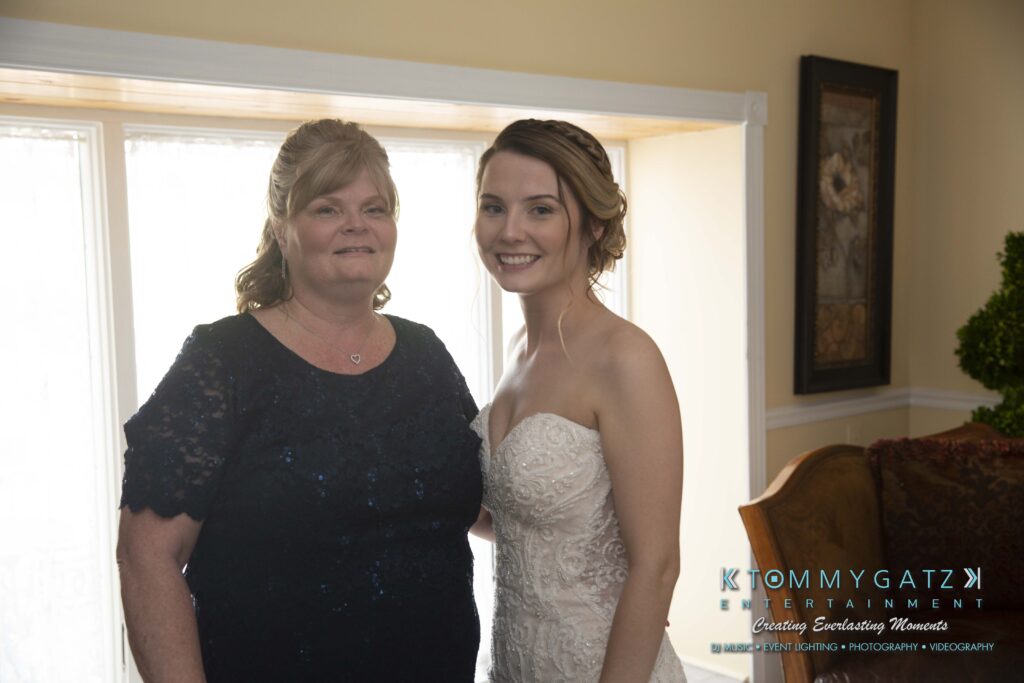 Two women standing indoors, one in a dark dress and the other in a white strapless gown. They are both smiling.
