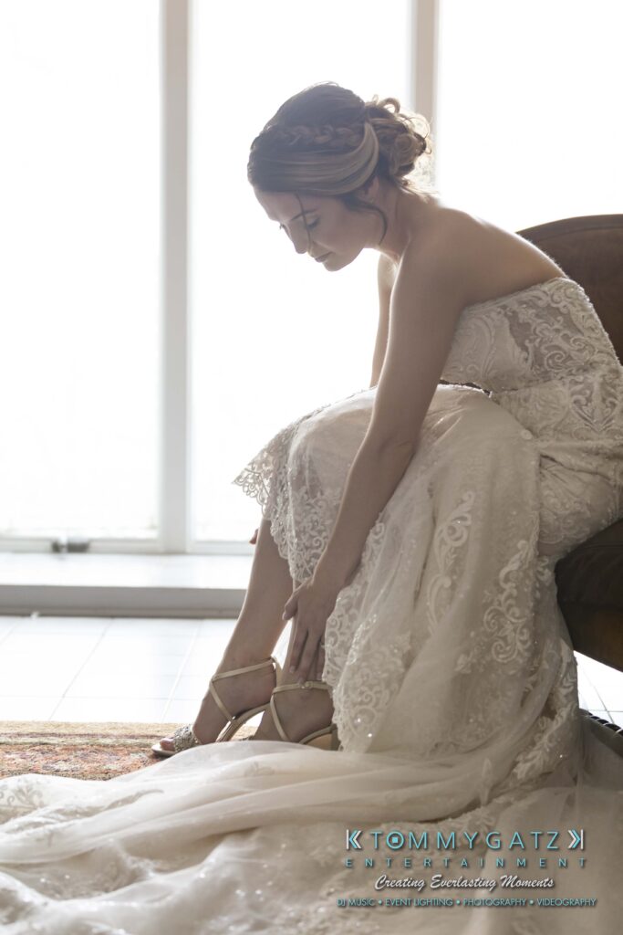 A bride in an elegant white lace gown sits on a chair, adjusting her high-heeled shoe by a large window.