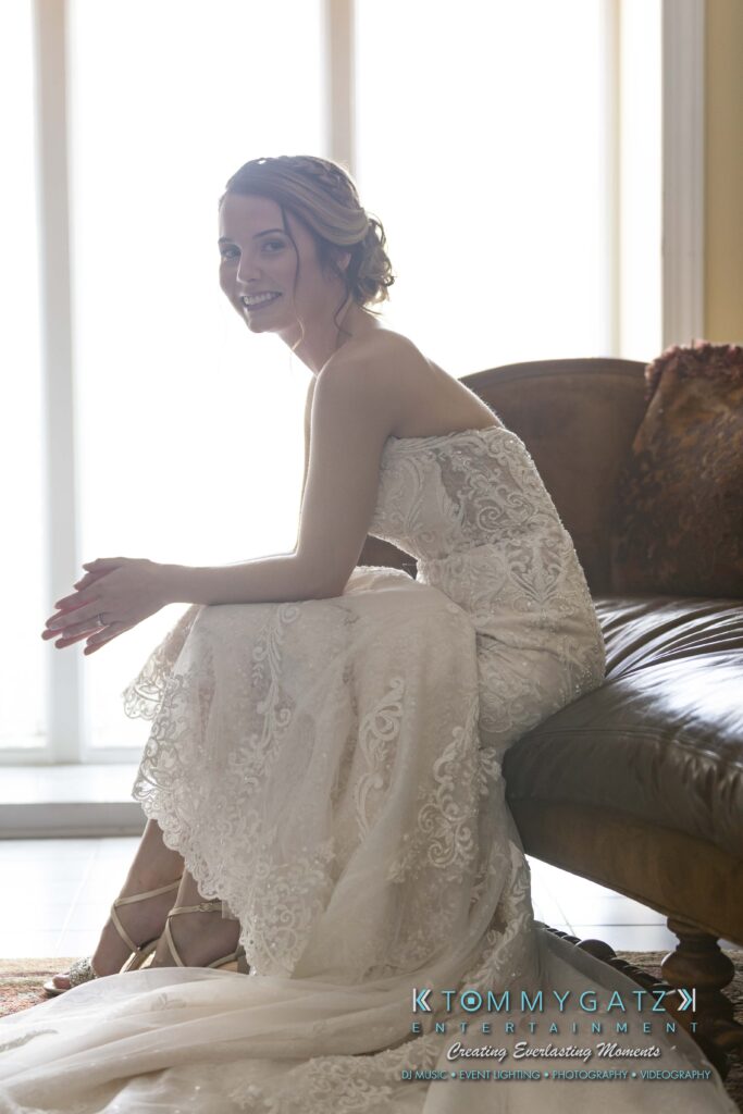 A bride in a lace strapless wedding dress sits by a window, smiling, with her hands resting on her knees.