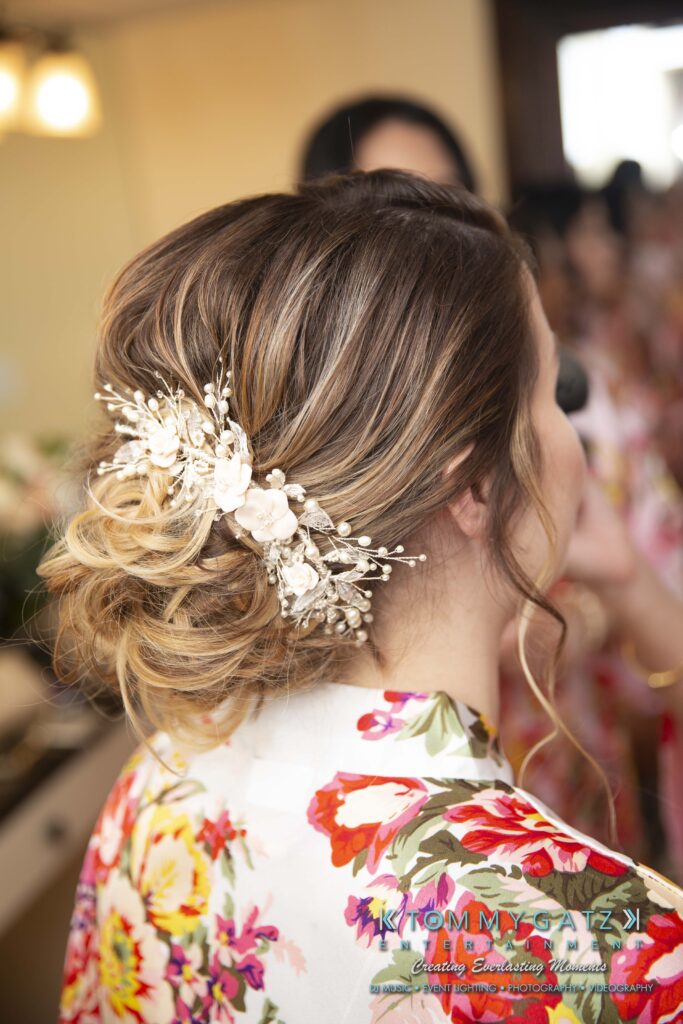 Woman with intricate braided updo adorned with white floral accessories, wearing a colorful floral robe, captured from behind.