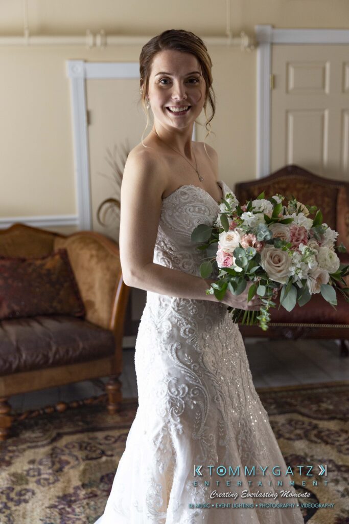 Bride in a white lace gown holding a floral bouquet, standing indoors on a patterned rug with a sofa and chairs in the background.