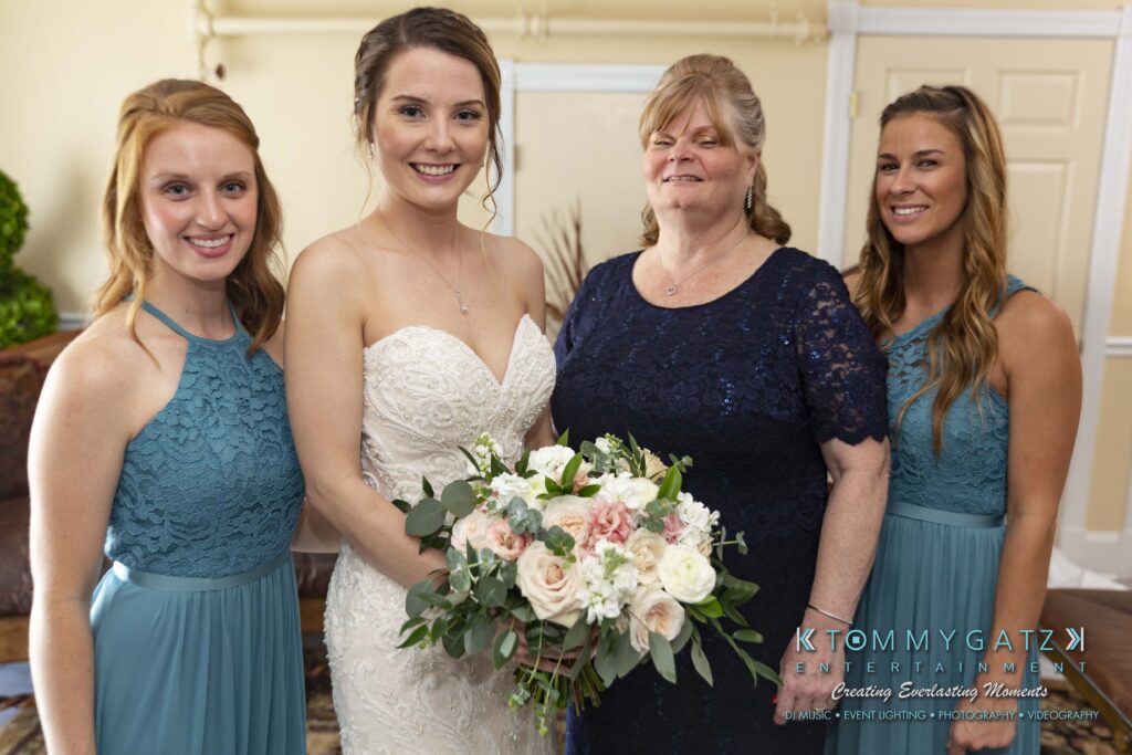 Bride in white dress holding bouquet, flanked by three women in teal dresses, standing indoors with neutral background.