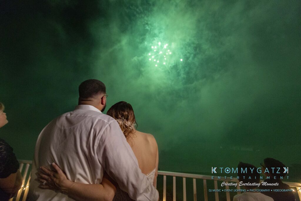 A couple embraces while watching fireworks in the night sky. They stand on a balcony with others, dressed formally.
