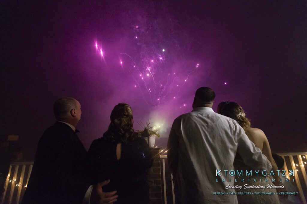 Four people in formal attire watch pink fireworks in the night sky from a balcony.