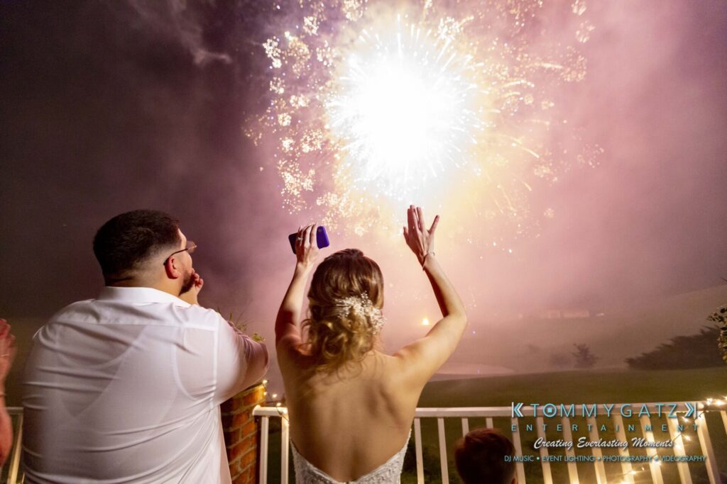 A couple watches fireworks at night, with the woman raising her hands in celebration.