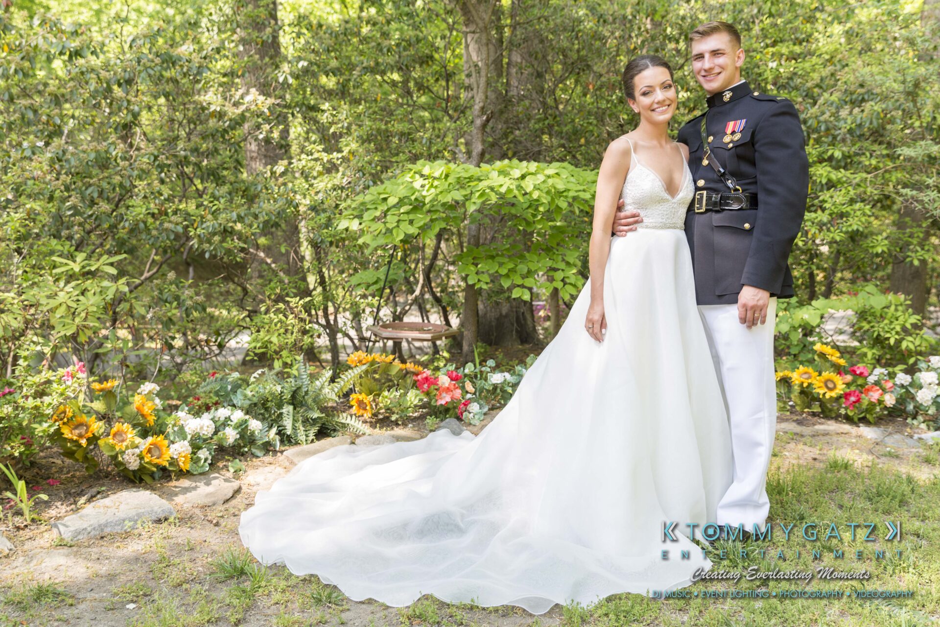 Bride in a white gown and groom in a military uniform stand smiling in a garden, surrounded by greenery and flowers.