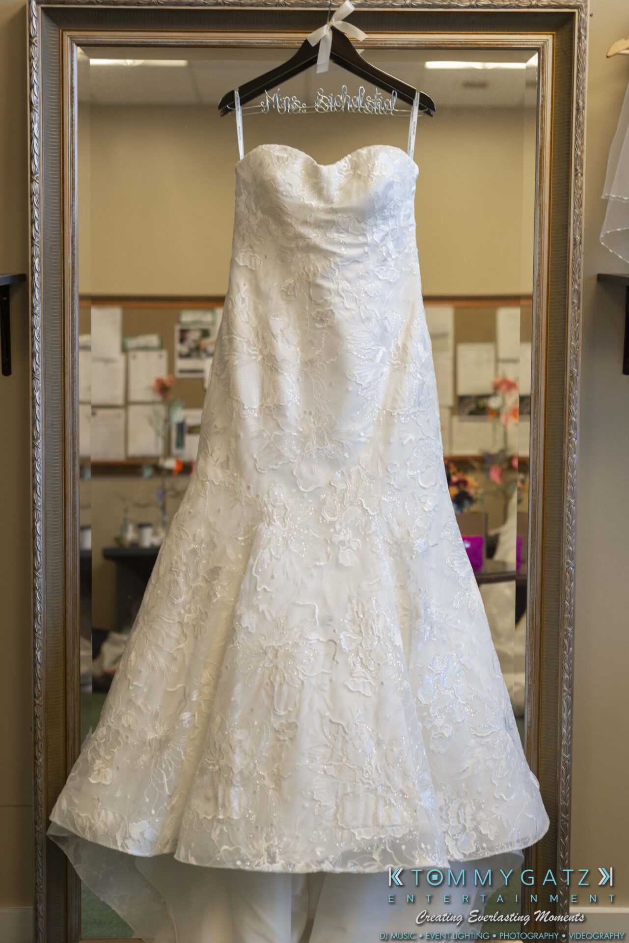 A strapless white lace wedding dress hangs on a wooden hanger in front of a large mirror.