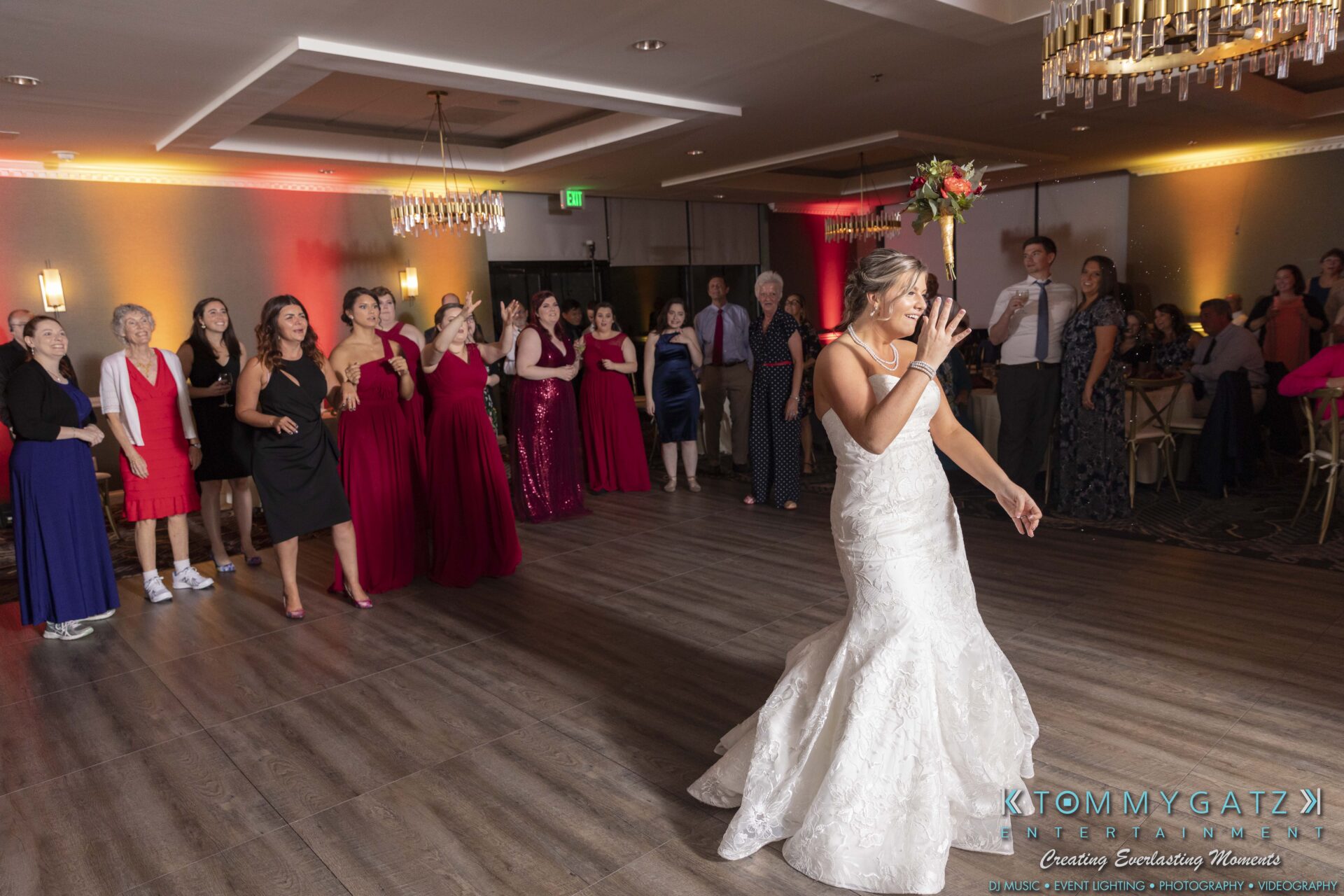 A bride in a white gown prepares to toss her bouquet in front of a group of women in a reception hall.