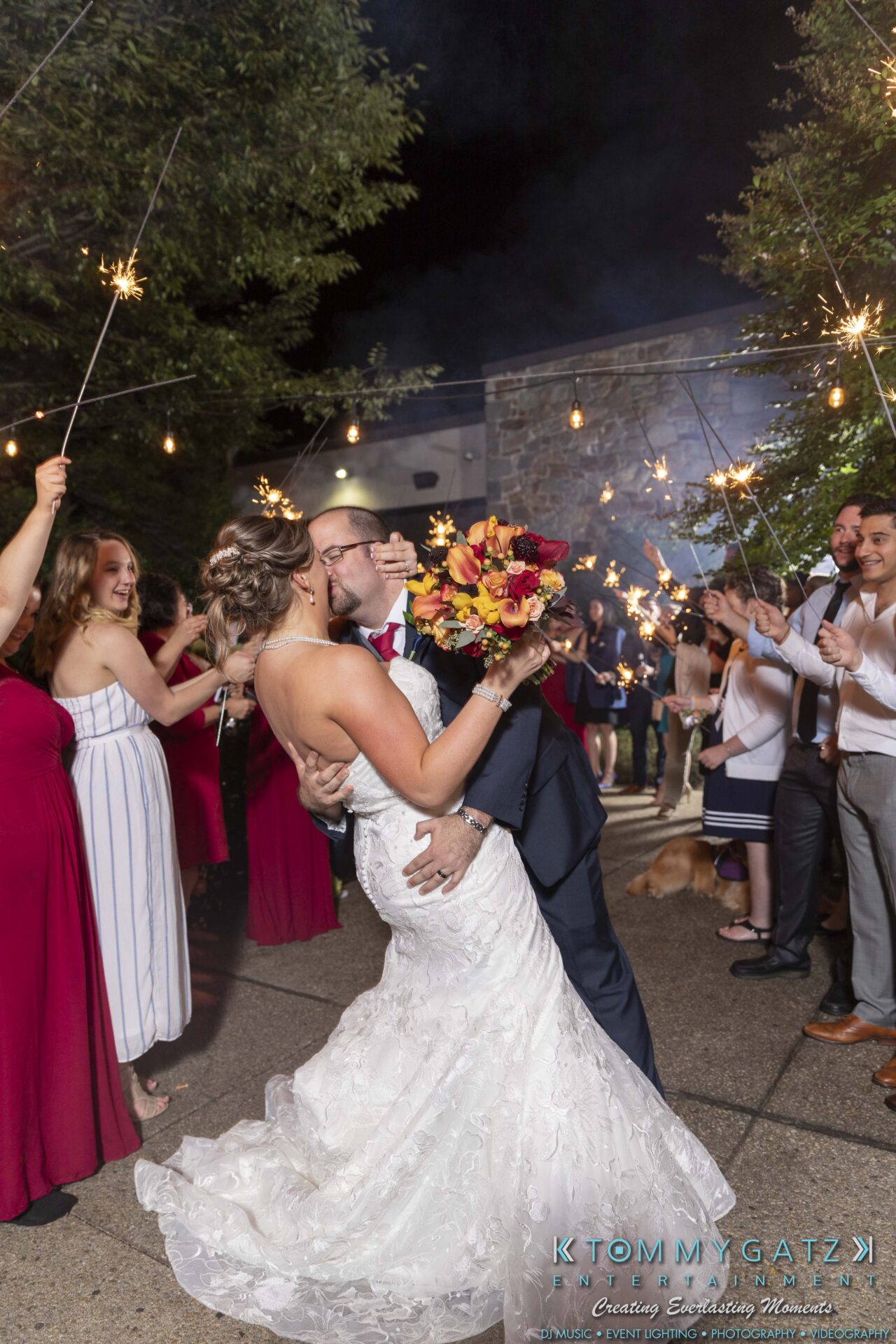A bride and groom kiss surrounded by guests holding sparklers. The bride holds a bouquet of red and yellow flowers.