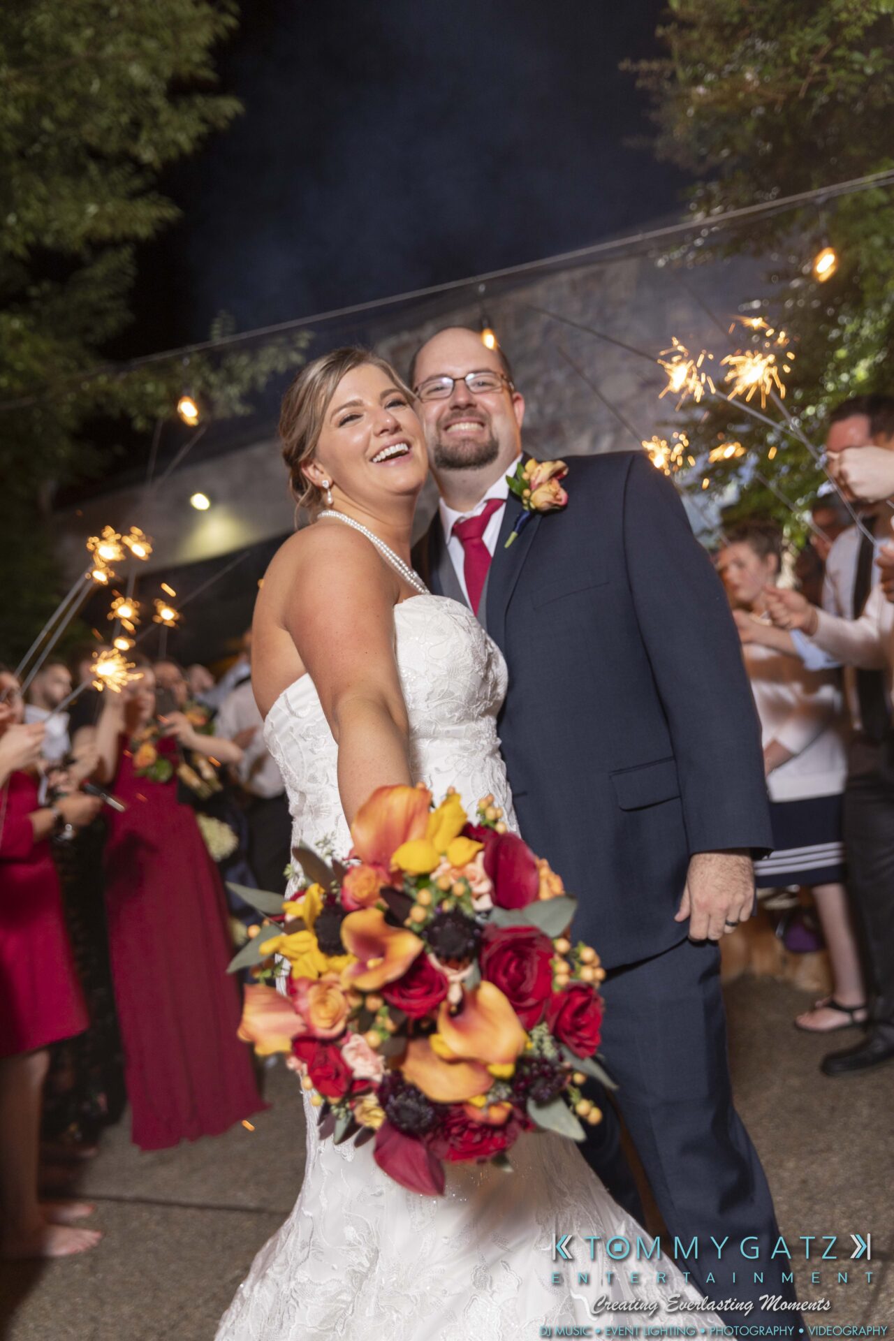 A bride and groom smile while holding bouquet, surrounded by guests with sparklers at night.