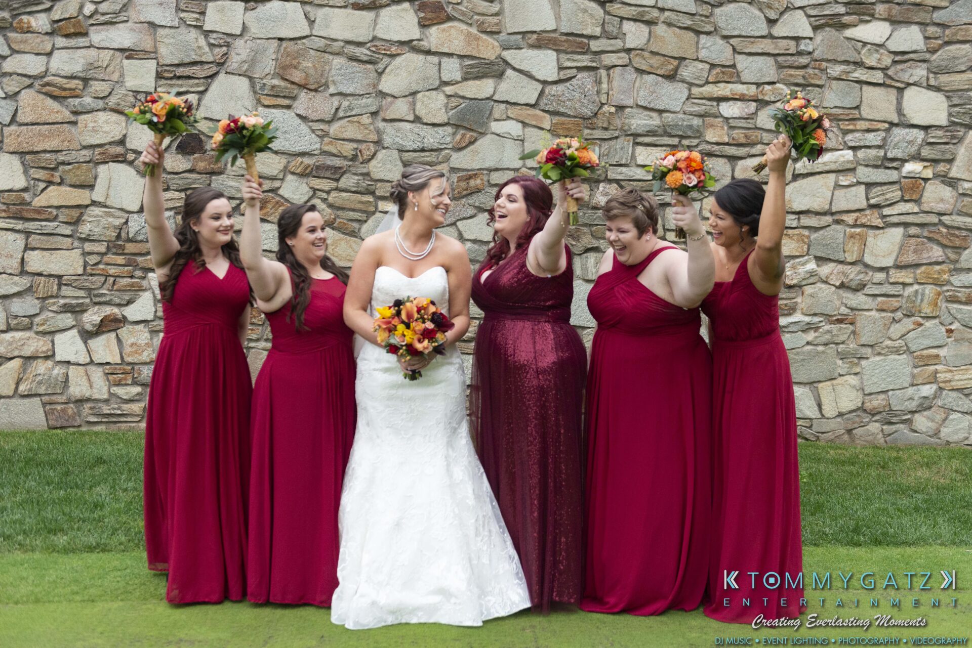 A bride in a white dress stands with five bridesmaids in red dresses holding bouquets, smiling in front of a stone wall.