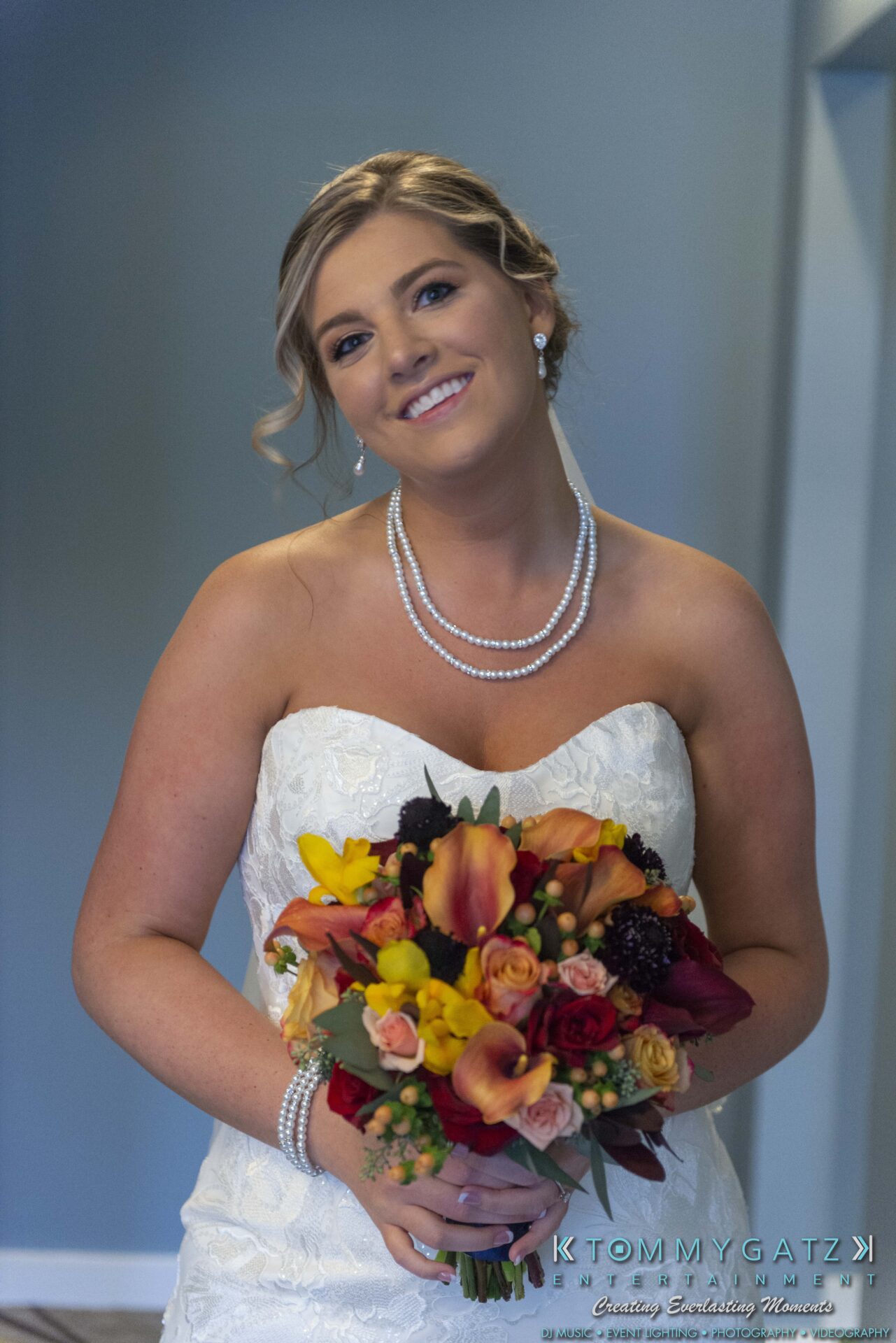 A bride in a strapless white dress holds a colorful bouquet. She is smiling and wearing a pearl necklace and earrings.