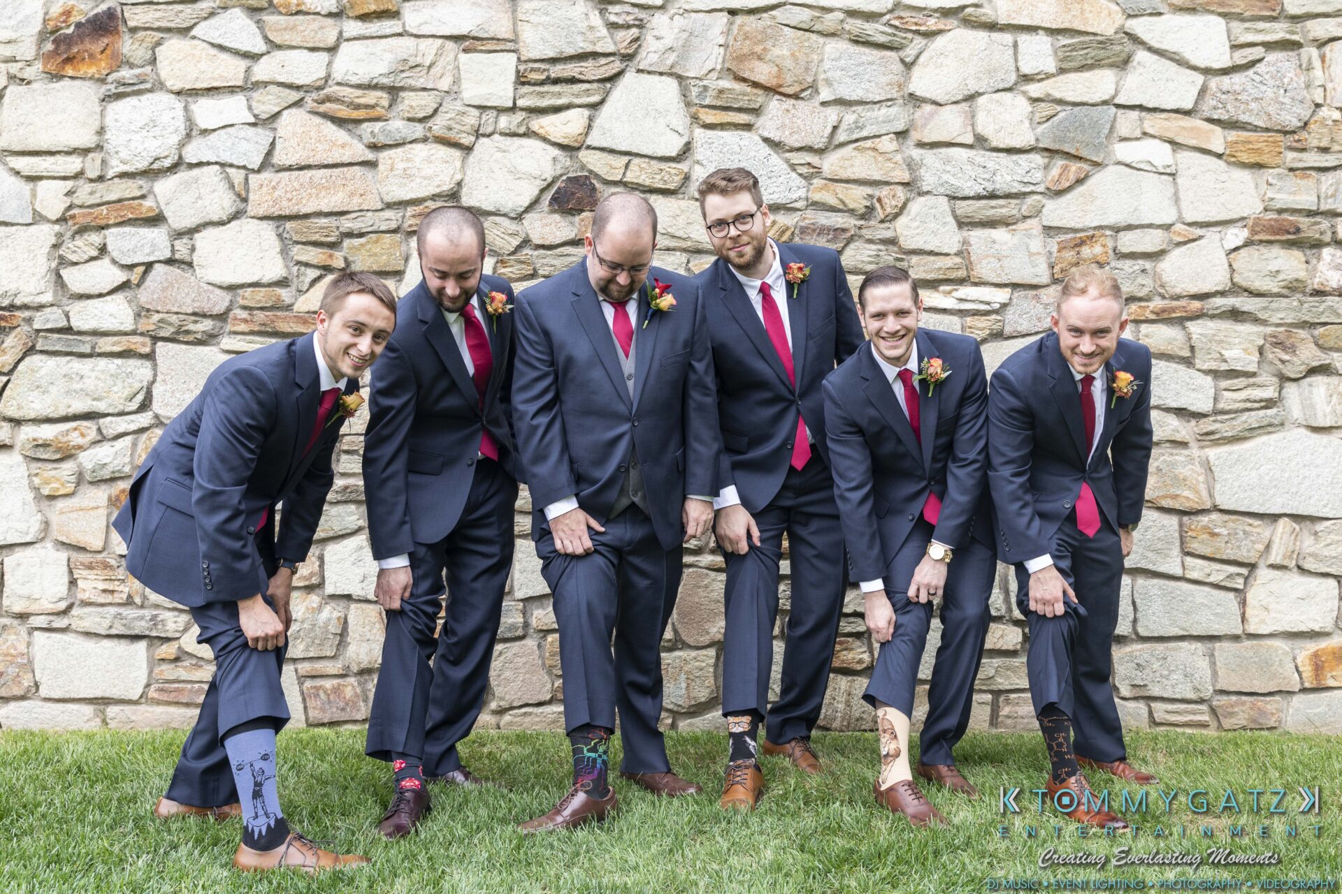Six men in suits stand outdoors, showing off colorful socks, against a stone wall backdrop.