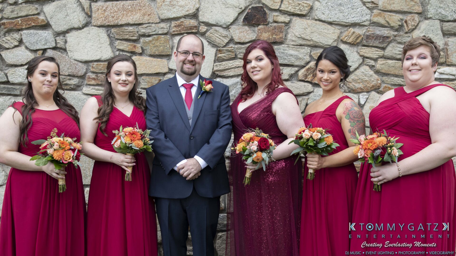 A group photo of a groom in a suit and five bridesmaids in red dresses holding bouquets, standing against a stone wall.